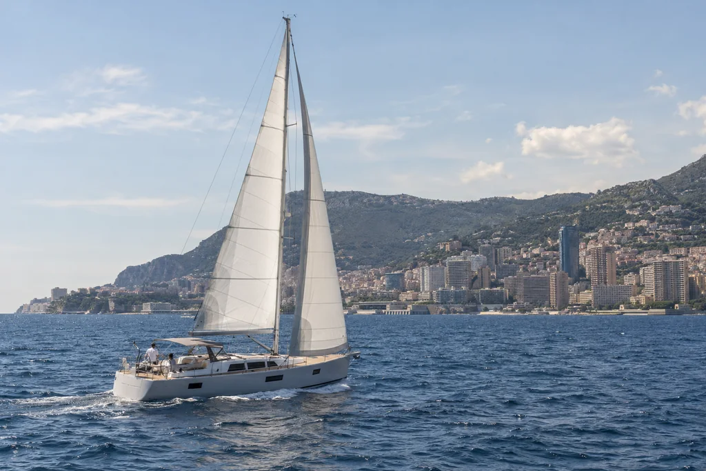 A slender unbranded sailing yacht under white sail on the Mediterranean, photographed from another vessel, Monaco coast far in the background